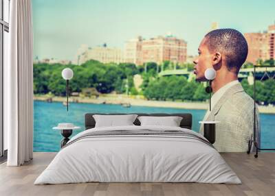 Young African American Man with short hair, traveling in New York City, standing by Hudson River in Manhattan under sun, looking, waiting, thinking. Green trees, buildings on background. Side View.. Wall mural