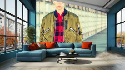 Young blonde American college student wearing green jacket unzipped, red patterned shirt, standing on walkway with glass wall on campus in New York, looking forward.. Wall mural