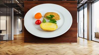 Slices of boiled potatoes, two tomatoes and a leaf of parsley, lie on a white round plate on a wooden brown table. Wall mural