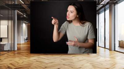 To be indignant and accuse the interlocutor, a young woman argues with the interlocutor by pointing her finger. A young brunette in a T-shirt is discussing with someone, portrait on black. Wall mural