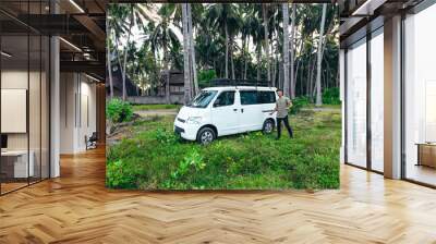 wide landscape of young man standing next to white camper van parked in tropical field with coconut trees at sunset in Bali Indonesia Wall mural