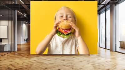 Cute little caucasian girl with blonde hair enjoying burger on a yellow background. Happy kid eating fast food burger Wall mural