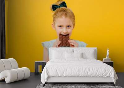 little girl tasting chocolate, look at the camera, in blue T-shirt, isolated yellow background, copy space Wall mural