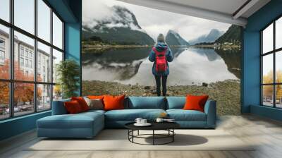 One woman stands in front of a lake mirroring mountains in blue jacket with red backpack Wall mural