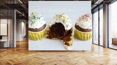 Two whole and one bitten chocolate cupcake in yellow cup with white whipped cream decorated with colorful sprinkles on white wooden table. Wall mural