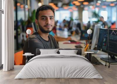 Confident young man with a beard smiling at the camera while sitting at his workstation in a vibrant and busy office setting, showcasing a creative and productive work atmosphere Wall mural