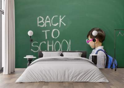 cute little boy preschooler is writing on blackboard with white chalk the words back to school.kid is wearing funny round party glasses with no glass inside.education,learning,classroom interior Wall mural