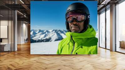 Man is posing at a ski resort, wearing a green jacket and black ski goggles, with snowy mountains in the background Wall mural