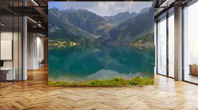 The green water of a mountain lake is reflecting the dramatic peaks surrounding it on a cloudy summer day Wall mural