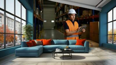 Warehouse worker in an orange vest and hard hat is using a digital tablet to check inventory in a large warehouse Wall mural