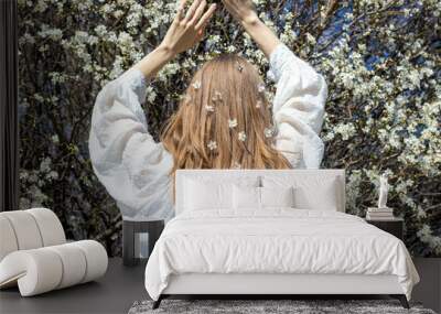 woman girl with long blonde curly hair against blossom cherry tree sun light blue sky spring is coming easter holiday.white little small flowers in female hair back view hands up touching branches Wall mural