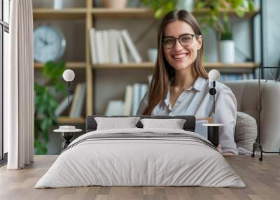 A smiling young female psychologist holds a clipboard while sitting in a modern office with bookshelves and greenery in the background. The setting is bright and welcoming, ideal for therapy sessions. Wall mural