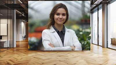 A young woman with a bright smile, dressed in a white shirt and apron, stands in a greenhouse surrounded by blooming flowers. She is holding a plant and appears confident and happy in her gardening bu Wall mural