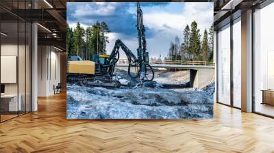 Side view at yellow drilling machine which stands on granite rock and boring holes for dynamite at rock blasting area - further rock exploding, new road construction. Evening shadows, green forest Wall mural