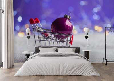 Crimson Christmas ball in small supermarket trolley on white knitted fabric with garland lights on purple bokeh background. Wall mural