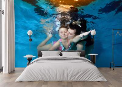 A smiling little girl is swimming underwater with her mother with her eyes open. Mom hugs and kisses her. They play sports. Portrait. Horizontal view Wall mural