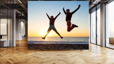 girl and guy jumping high with arms up spectacular sunrise at ocean coast Wall mural