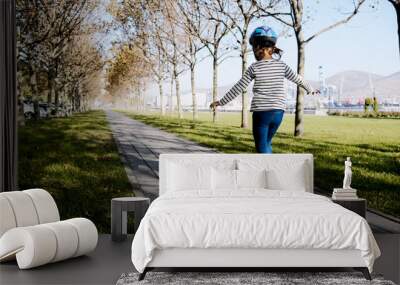 Little girl is slowly skateboarding in protective helmet in the park on path at sunny autumn weather. Wall mural