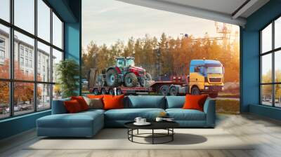 The semi truck is hauling farm equipment on a flatbed. The heavy industrial truck semi trailer flatbed platform transport one big modern farming tractor machine on the road. Evening sun. Wall mural