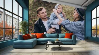 Stylish family in the autumn forest. A guy in a leather jacket and a young girl in a gray-blue dress play with his son in a meadow of a pine forest in the mountains against the background of a fence  Wall mural