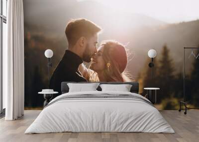 Stylish model couple in the autumn mountains. A young guy and a girl hug and kiss on a background of a forest and mountain peaks at sunset. Wall mural