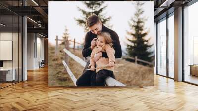 Stylish young family in the autumn mountains. Dad and daughter are sitting on the fence and hugging against the background of the forest and mountain peaks at sunset. Wall mural