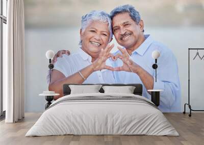 A happy senior couple with a heart sign with their fingers and enjoying fresh nature air on vacation at beach while bonding. Portrait of retired couple hugging at beach with smile and love together Wall mural