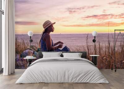 Black woman, relax and book on beach in sunset with diary or journal for vacation in nature outdoors. African female person relaxing on ocean coast with notebook in morning sunrise and holiday travel Wall mural