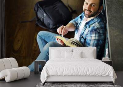 Portrait of joyful young man reading book in his living room.Student holding and reading book. Wall mural