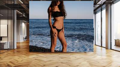 sexy girl brunette in a black swimsuit with a slender figure posing on the beach by the sea on vacation Wall mural