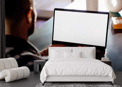 Over shoulder view of a young business man using computer laptop in front of an blank white computer screen in his home living room. Photo ready for mockup. Wall mural