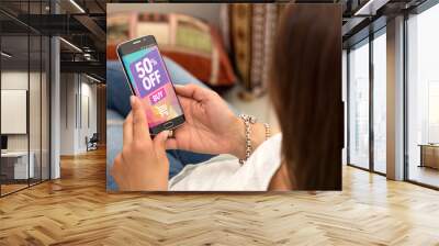 Promotion, special offer in a cell phone. One young woman in her home resting in armchair holding a smartphone with a advertising 50% off  in the screen. Wall mural