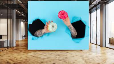 The wrists of a man and a woman came out through a torn hole in the blue paper holding a donut. Wall mural