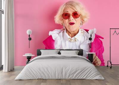 amazed elderly female in stylish clothes posing at camera isolated on pink background, surprised emotional lady in sunglasses Wall mural