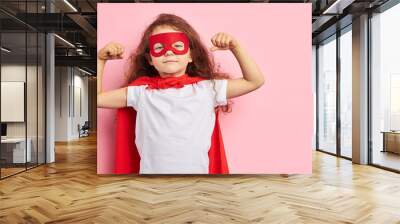 attractive little curly girl wearing red hero suit and mask showing how she is strong isolated over pink background Wall mural