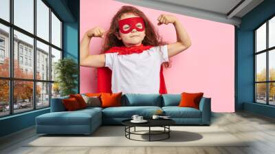 Beautiful little curly girl wearing red hero suit and mask showing how she is strong isolated over pink background Wall mural