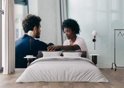 Caucasian boyfriend and his african girlfriend practice yoga, sit crossed legs, holding hands together, looking at each other while sitting on wooden floor in spacious room. Wall mural