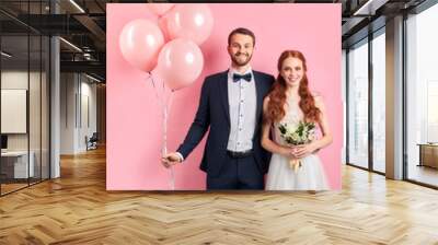 caucasian wedding couple posing together isolated over pink background. Handsome man in tuxedo holding pink air ballons, woman in white wedding dress holding bouquet of flowers Wall mural