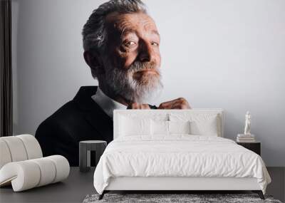 Confident old-aged gentleman in tux fixing his bowtie with both hands and looking at camera while standing against white background. Wall mural