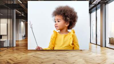 Cute african american girl wth curly fluffy hair holding magic wand, isolated on white studio wall. Funny little black girl wearing yellow casual shirt. Childhood lifestyle concept Wall mural