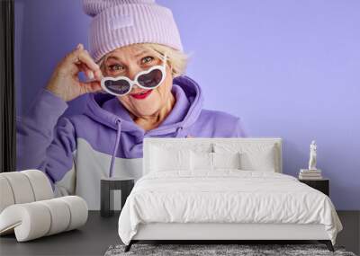 elderly woman holding hands on chest smiles at camera isolated over purple background, fancy female in pullover, hat and sunglasses Wall mural