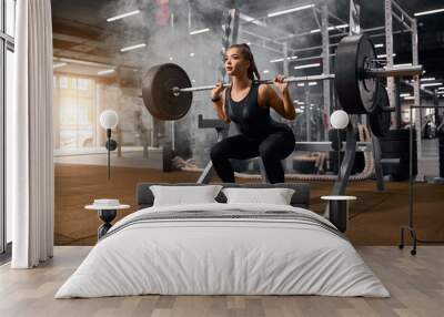 Female weightlifter is getting ready to stand with heavy barbell, squatting with weight, looking away with pleasant expression, enjoying training in gym, portrait, side shot Wall mural