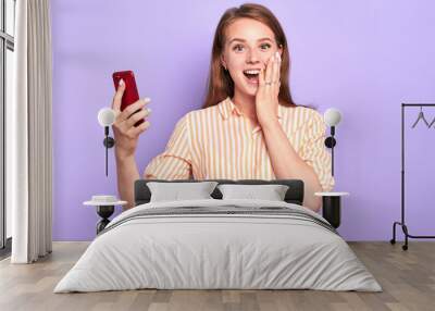 Front view of suprised funny young woman smiling broadly, holding red cell phone, cant believe in her success, happy and emotional, dressed in stylish shirt, standing at light purple wall. Wall mural