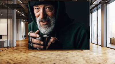 Frozen homeless bearded old man with grey hair and wrinkled face, looks at camera with grateful expression and holds a steel mug with coins given as alms, posing at studio over black background Wall mural