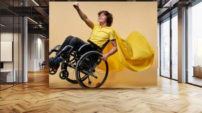 handicapped man in yellow cloak pretend to be superhero, raising hands up. isolated in studio on beige background. be happy and strong despite life's difficulties Wall mural