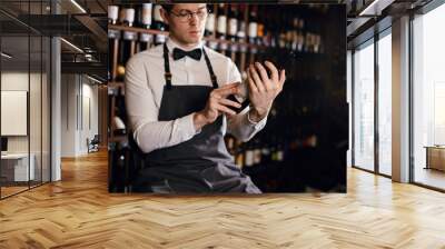 Handsome and young smiling brown haired sommelier with a bottle of wine on the background of dark wine house with shelves of bottles with alcohol Wall mural