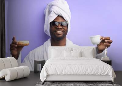 Handsome attractive man in towel and bathrobe holding cup of tea and donut, emotional. Cool boy in eyeglasses posing with donuts isolated over purple background Wall mural