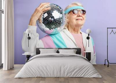 happy grandmother in sports clothes looking at camera with a pleasant smile while posing with disco ball in studio over purple background Wall mural