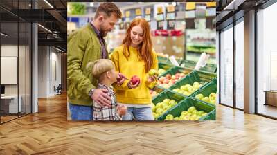 husband and wife with a kid buy fruits, apples. family of three choosing fresh apple in fruits department of supermarket or market Wall mural