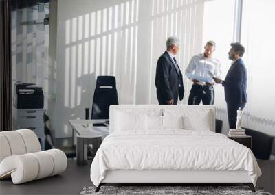 International male colleagues dressed in formal business suits and one dressed as Arab, stand near big windows talking, sharing impressions after corporate seminar Wall mural